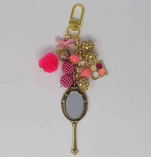 Get Ready Glam purse charm featuring a gold hand mirror charm, a miniature eyeshadow palette charm, a fluffy hot pink pompom, and a cluster of pink and gold faceted beads.