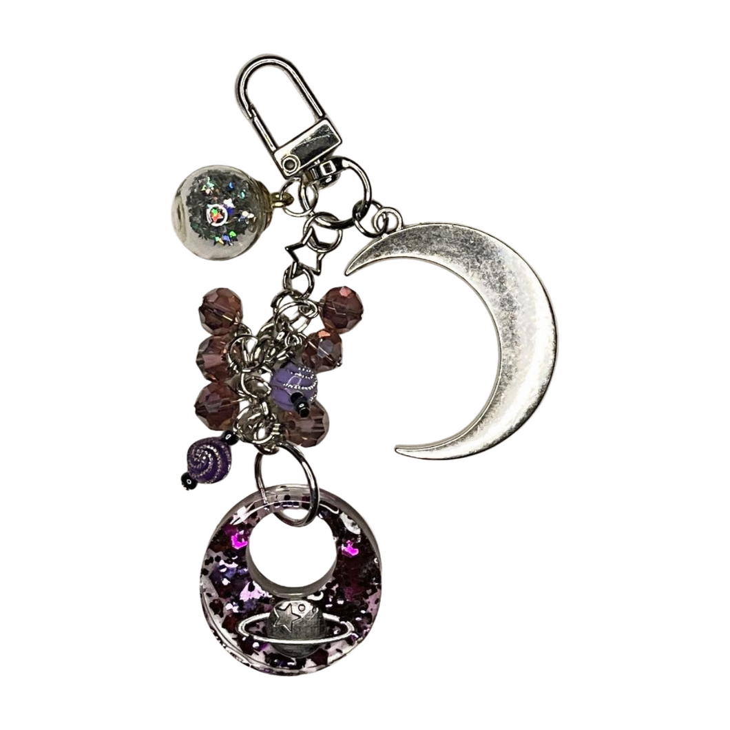 Celestial-themed purse charm featuring a silver crescent moon, a glittery resin pendant with a planet design, purple beads, and a star accent, attached to a keyring clip.