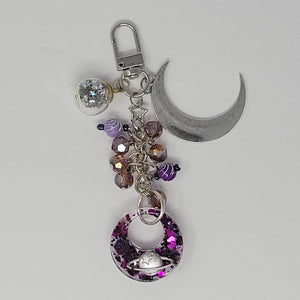 Celestial-themed purse charm featuring a silver crescent moon, a glittery resin pendant with a planet design, purple beads, and a star accent, attached to a keyring clip.