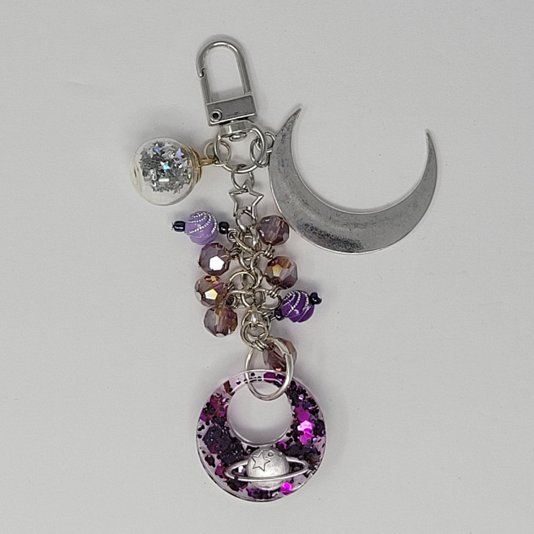 Celestial-themed purse charm featuring a silver crescent moon, a glittery resin pendant with a planet design, purple beads, and a star accent, attached to a keyring clip.