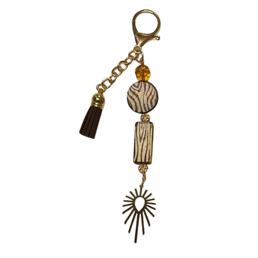 Wild Radiance purse charm featuring a gold clasp, a brown tassel, a zebra-print bead, a rectangular zebra-patterned bead, an amber-colored bead, and a gold sunburst charm, designed for stylish accessorizing of bags or purses.